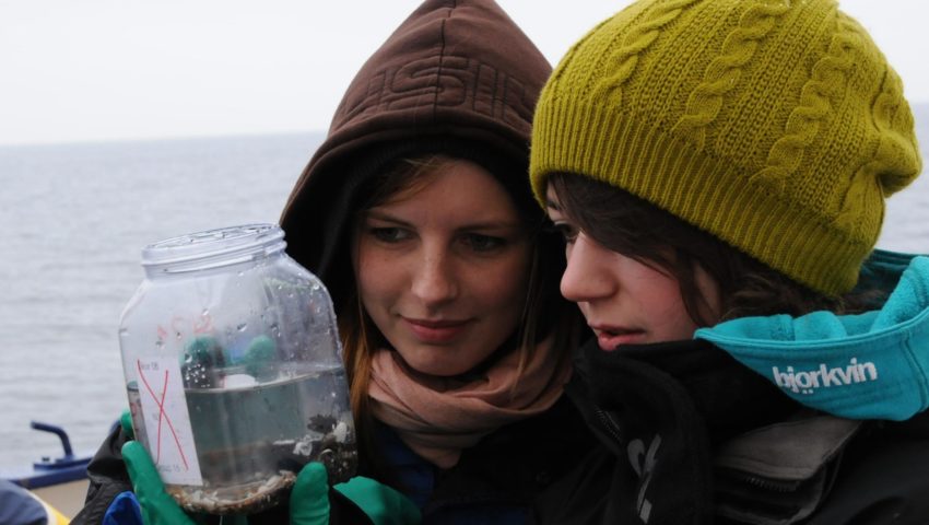 Zwei Schülerinnen stehen bei einer Exkursion am Meer und sehen sich eine wissenschaftliche Probe an