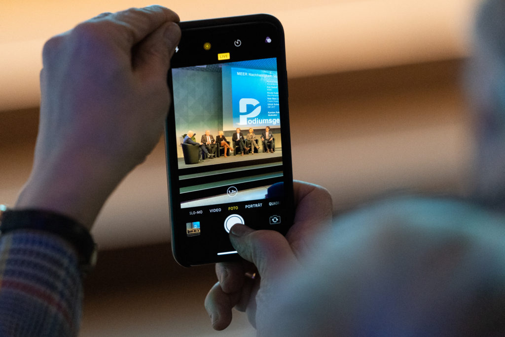 Aufnahme eines Smartphones mit einer Aufnahme der Podiumsdiskussion auf der Bühne bei der Auftaktveranstaltung der DAM