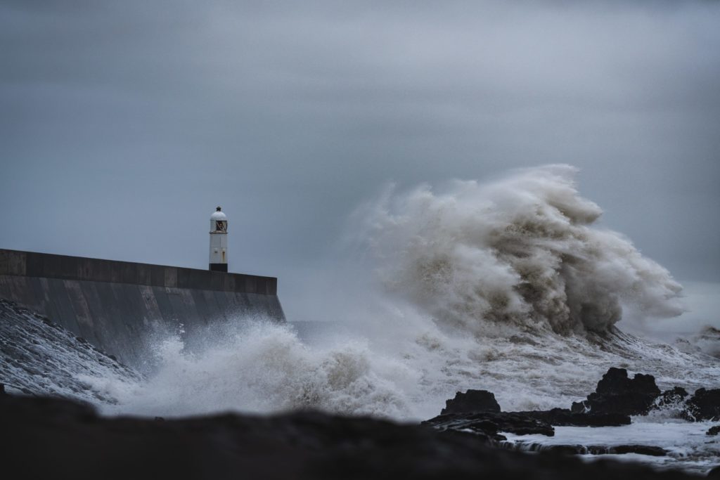 Research: Eine große Welle trifft mit voller Wucht auf eine Kaimauer mit Leuchtturm bei Sturm.