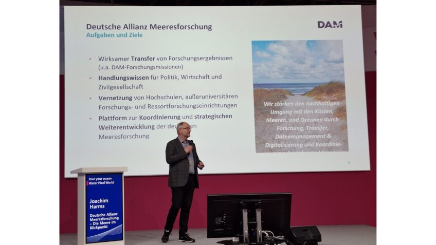 Dr. Joachim Harms gives a lecture on a stage at BOOT 24, behind him a presentation with key points on the work of the DAM